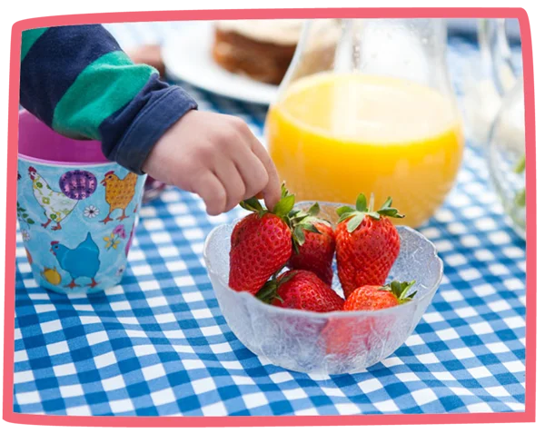 Small child taking strawberries from a bowl that also has a jug of orange juice.