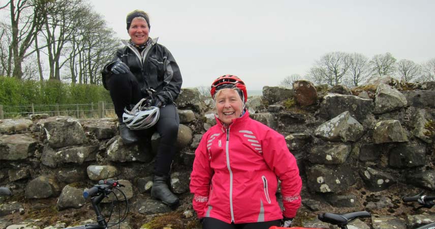 Two women in cycling wear posing in front of a stone wall on a rainy day