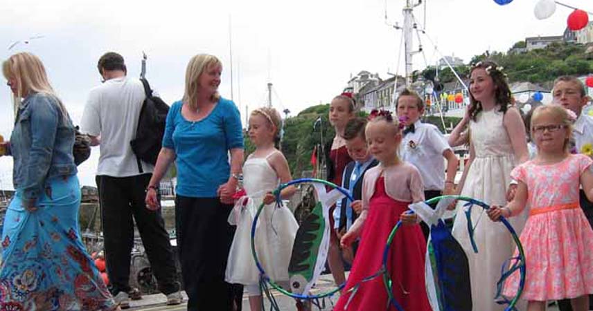 Festival in Cornwall with children dressed smartly holding hoops. A boat is in the background, and bunting and balloons can be seen in the distance.