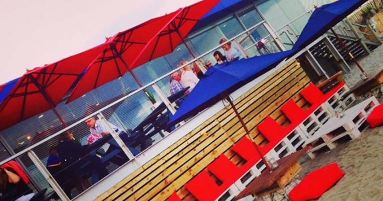 Skewed image of cafe on the beach with customers eating at benches under coloured umbrellas.