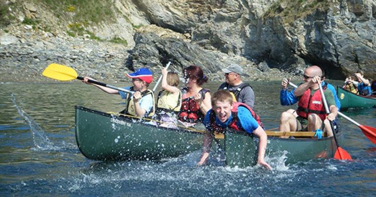 Three canoes full of adults and children canoeing down a river in Cornwall. Two young boys in opposite canoes are splashing each other.