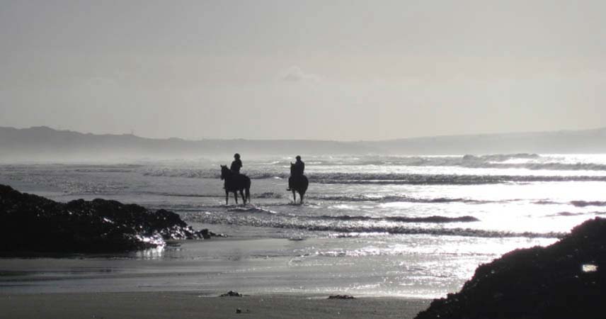 Two people horse riding at sunset on a beach in Cornwall.