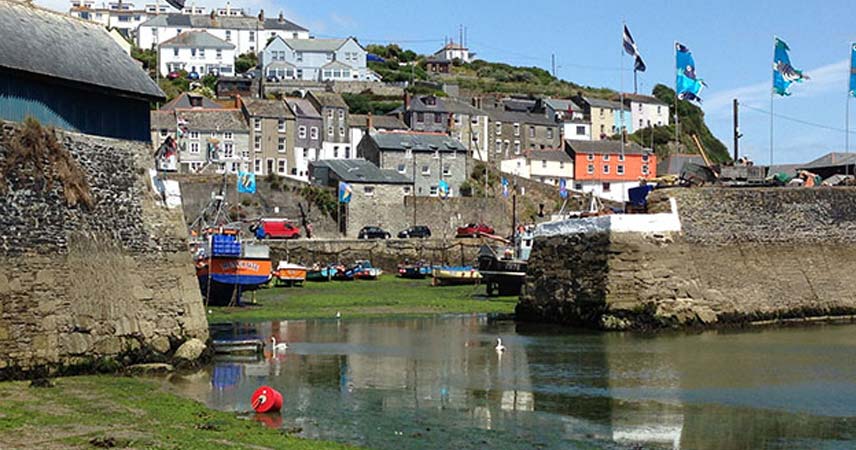 Entrance to Mevagissey harbour in Cornwall with blue flags flying and houses on the hill.