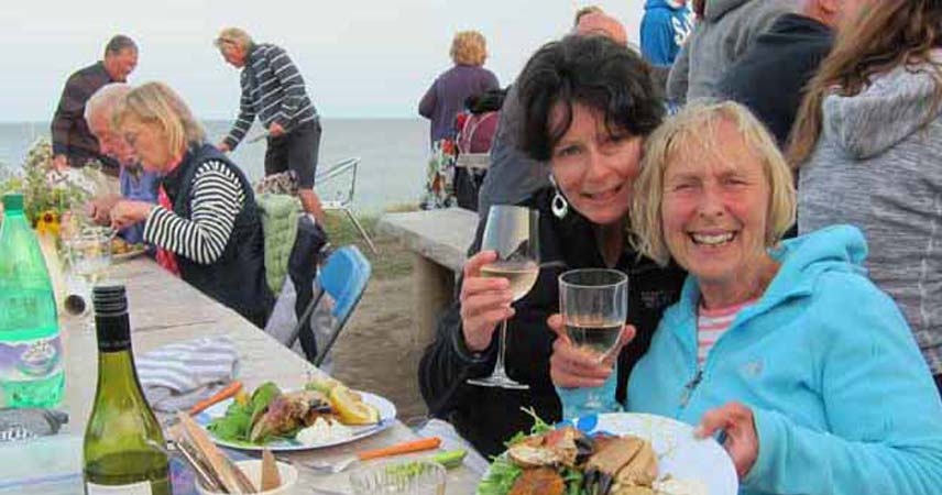 Groups of people eating on tables by the sea in Cornwall, with two people smiling for the camera with glasses of wine.