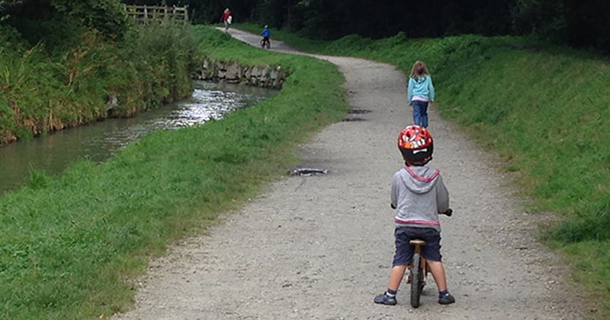 Child riding a bike by a river in Cornwall along a path, with two other children in front of him.