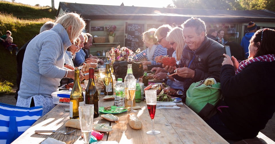 Large group of people eating together outside on a long dining table surrounded with a few bottles of wine.