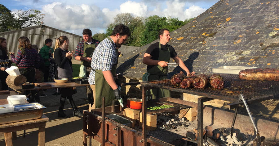 Two men grilling meat at Nancarrow Farm for a group of people.