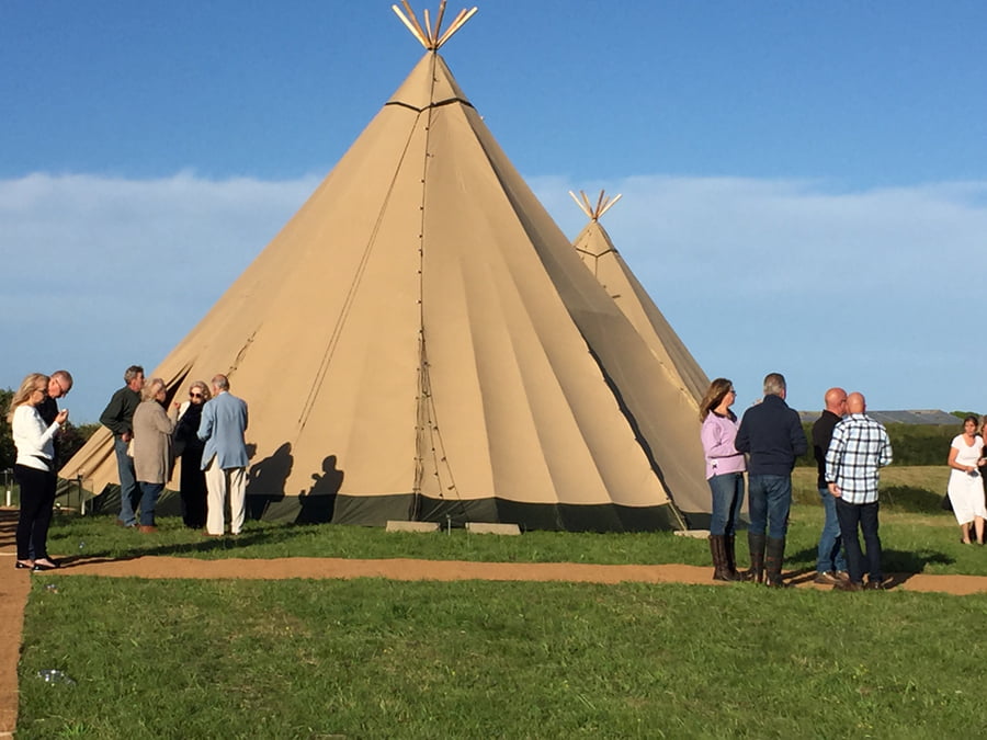 Two large tipis in a field on a sunny day, with groups of people talking around them.