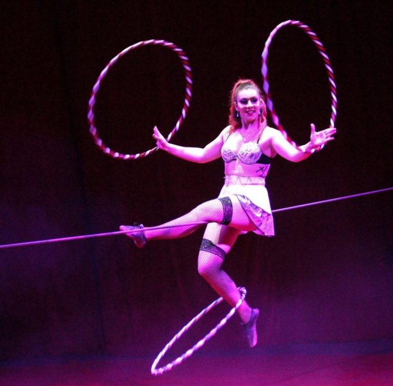 Female circus performer spinning three hoola hoops as she sits on a high wire.
