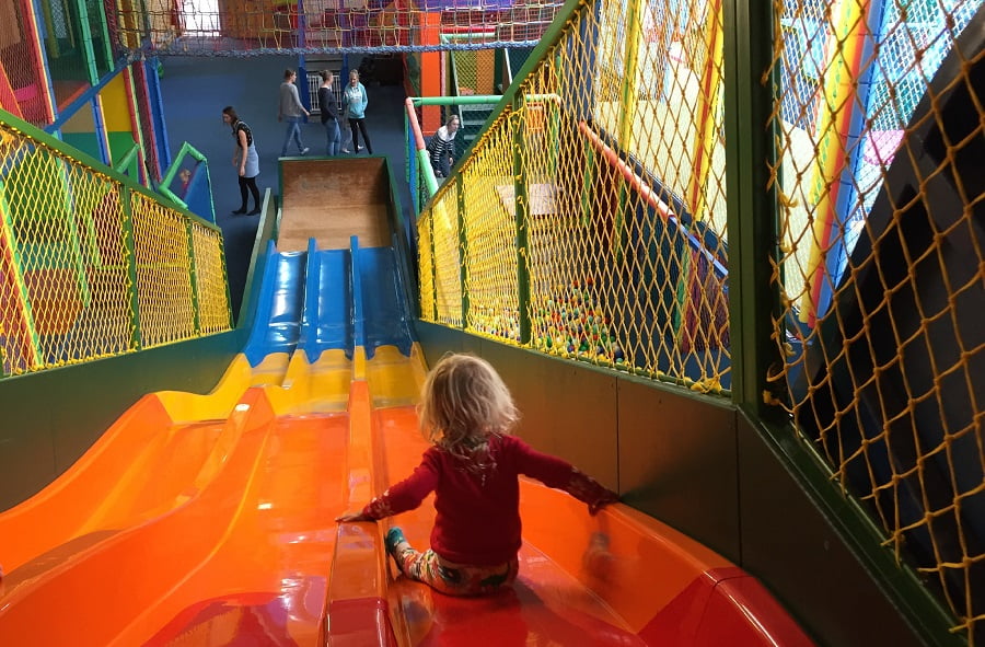 A child slides down the slide in an indoor play area, with four adults waiting at the bottom.