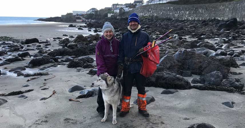 Nanny Pat with man and husky on a rocky beach holding litter picking tools.