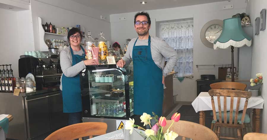 Smiling man and woman in blue aprons stood in a coffee shop