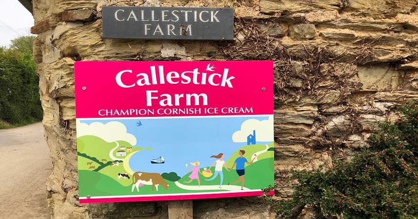 Callestick Farm pink sign on a stone wall, with illustration of family and animals on the coastline.