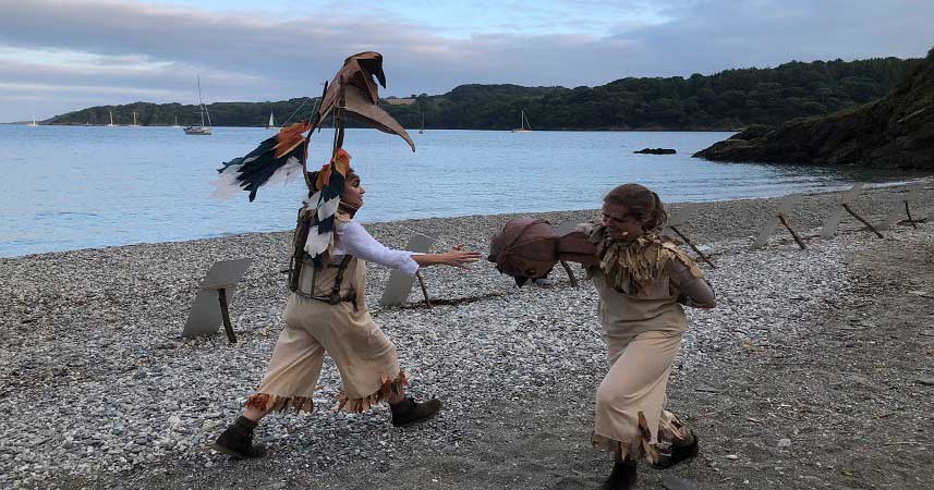 Two actors with bird puppets on a beach at dusk