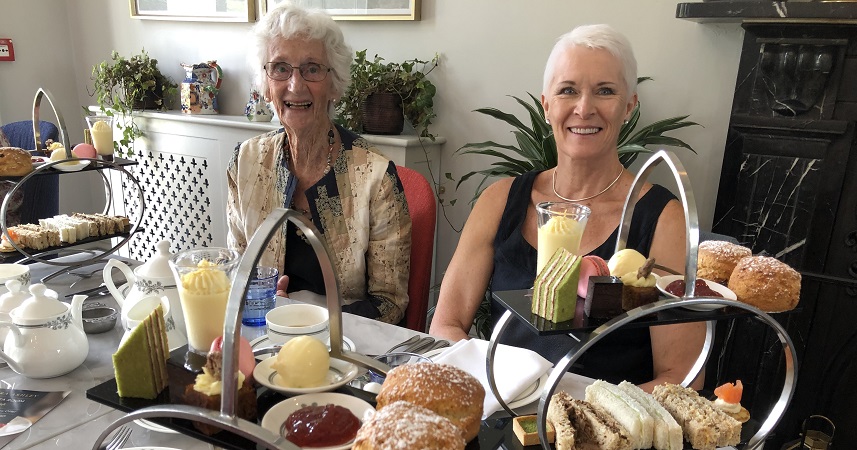 Two well dressed ladies sat at a table with afternoon tea stands, including small cakes, scones and sandwiches