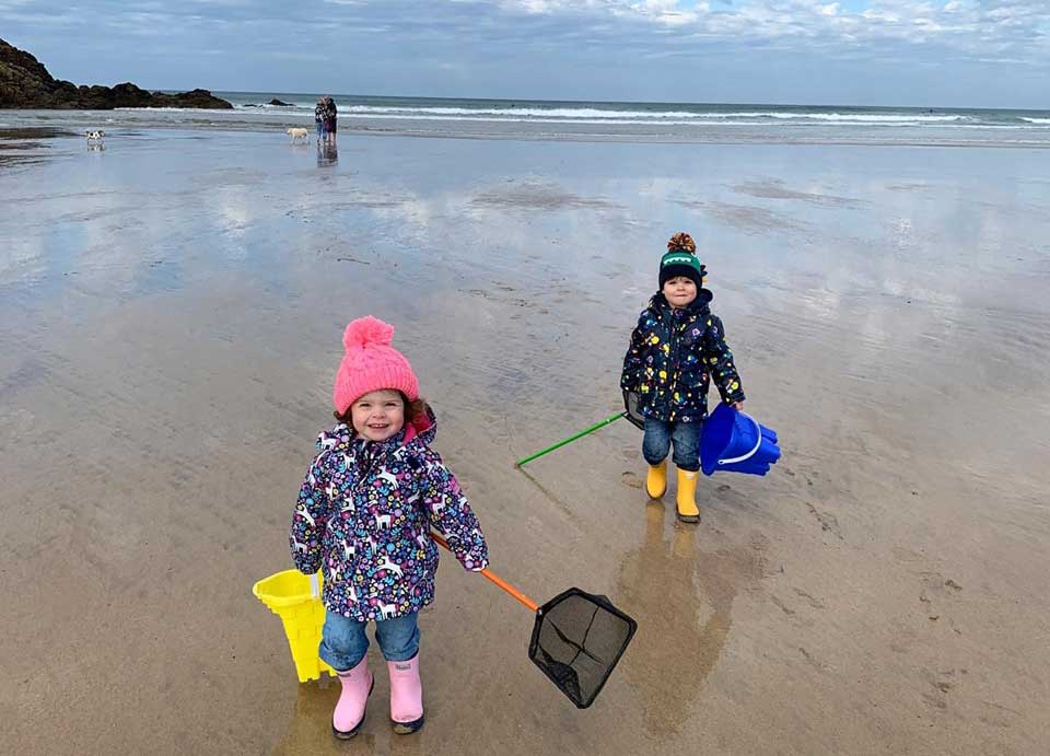 Two small children on a cornish beach with woolie hats, holding buckets and nets