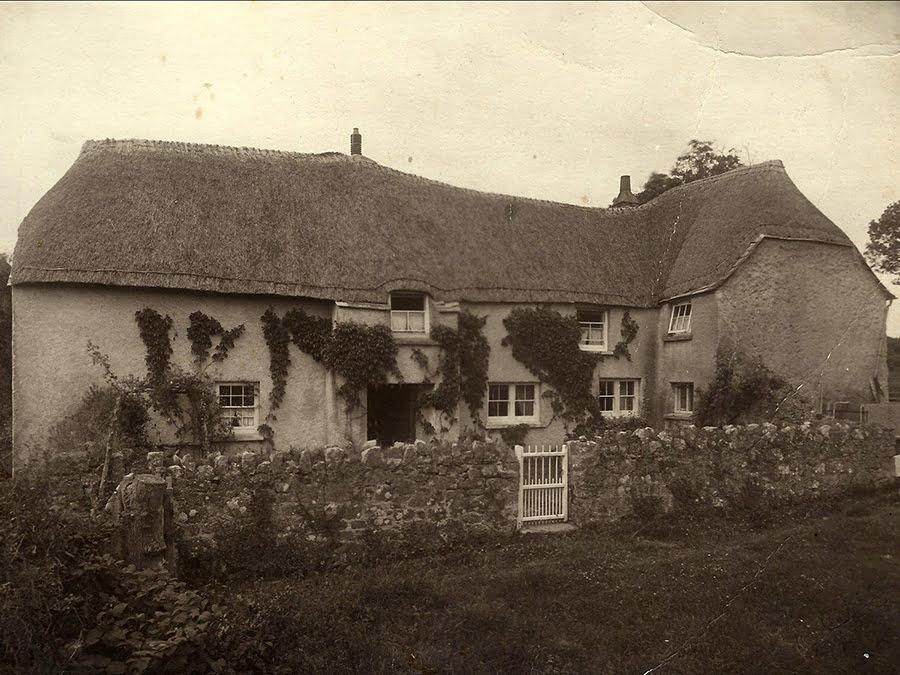 A black and white image showing a traditional thatched Cornish farmhouse with a stone wall and white gate