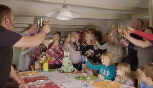 Group of guests raising glasses and celebrating with a large green birthday cake. There are place mats and party poppers on the table.
