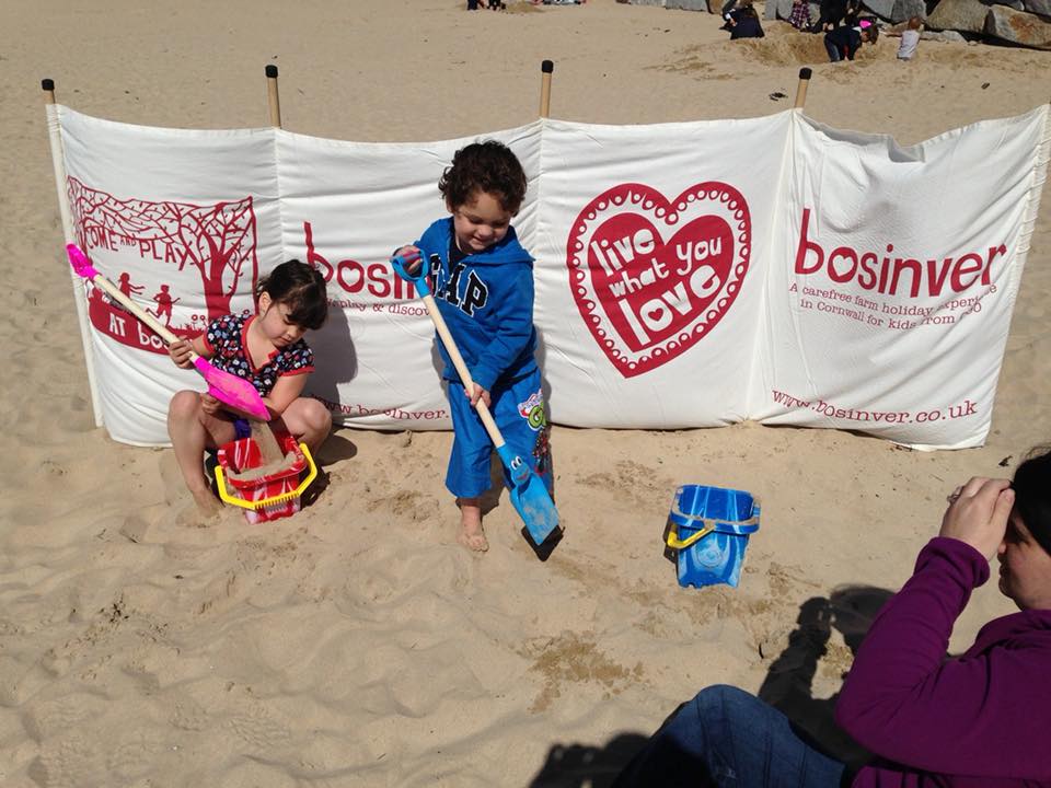 2 younge children on beach with buckets and spades in front of a windbreak.