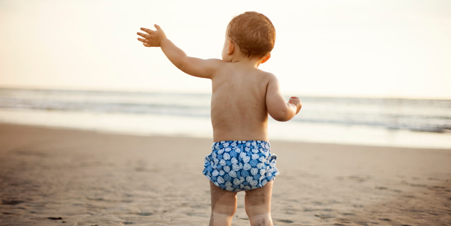 baby boy stood on the beach looking out to sea wearing swim shorts with one arm in the air like he is waving