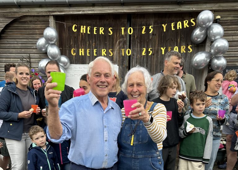 Couple at the front of a group of people raising a glass in celebration. Banners and balloons in the background.