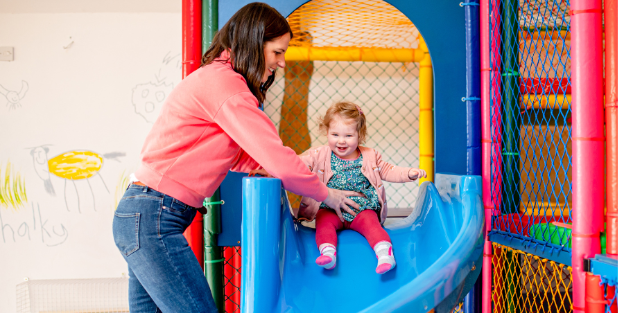 mother holding daughter at the top of blue slide in soft play area