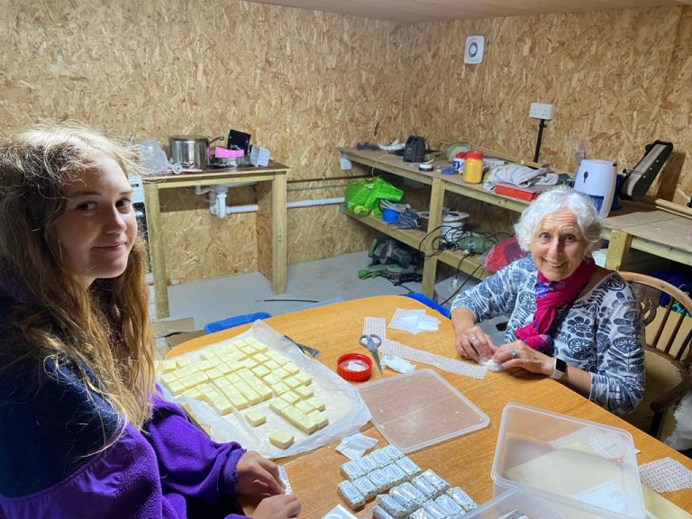Young lady and older lady sat around large table in workshop wrapping homemade soaps