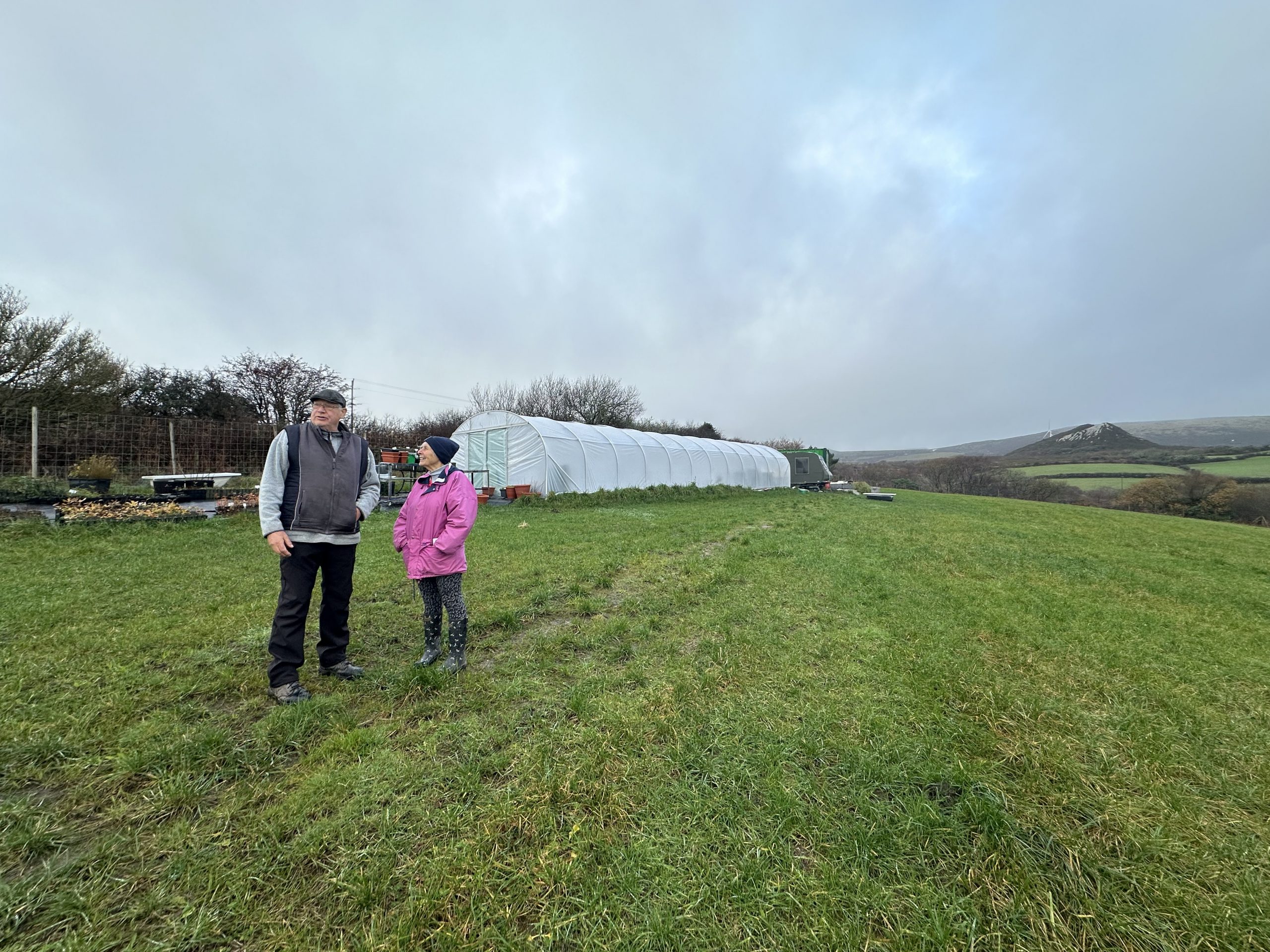 A ladty and gent in a stood in a community tree nursery with poly tunnel in the background